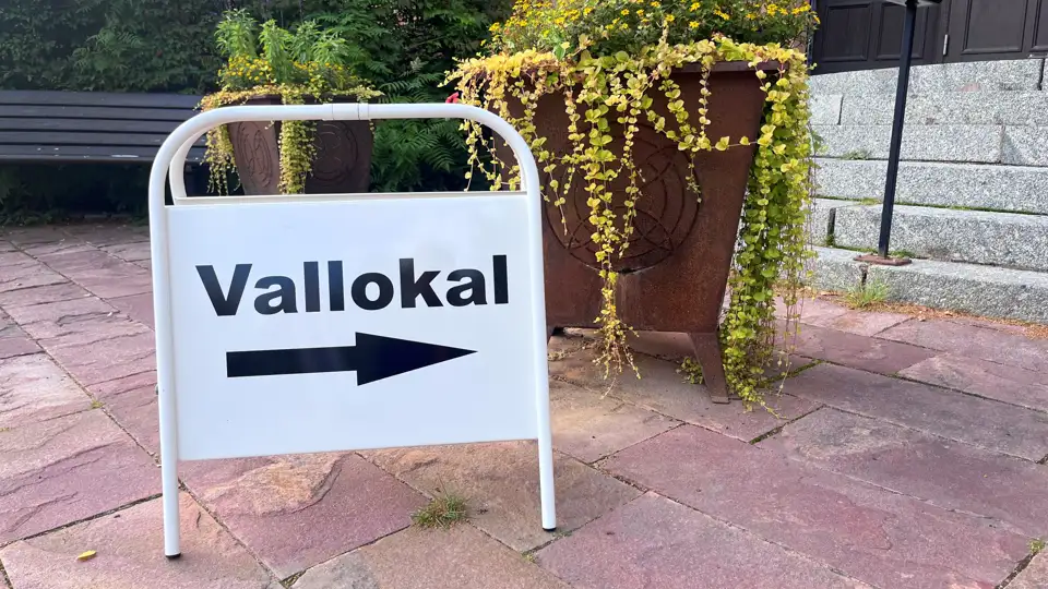 En vit skylt med texten Vallokal som står på trottoaren bredvid en krukväxt.