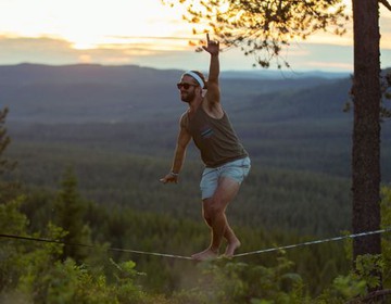 En muskulös man balanserar på en lina spänd mellan två träd på Orsa finnmark. Foto.
