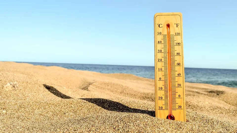 Liten inomhustermometer står uppställd på en sandstrand. Det är en väldigt varm sommardag. Foto.